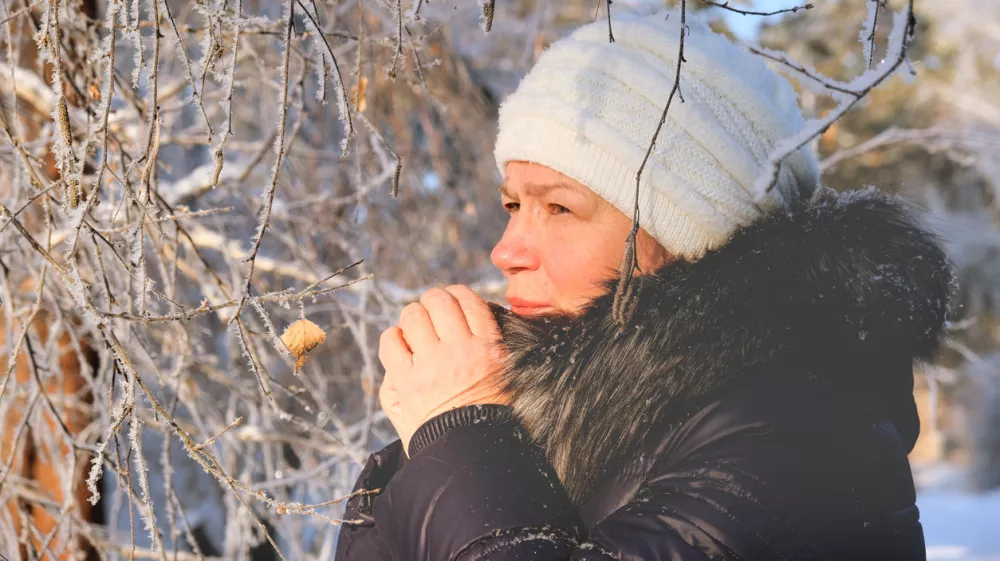 Woman in the blue padded jacket and white cap under snowfall at the street. Pensive attractive mature woman with a quiet smile. Sunny winter vacation outdoors. Deep breathing freshness