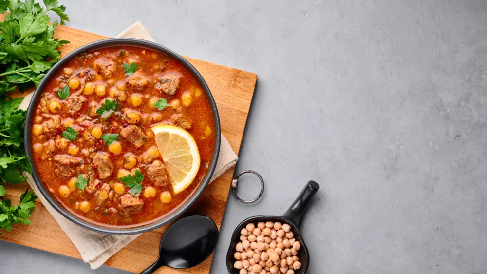 Moroccan Harira Soup in black bowl at grey concrete table top. Harira is Moroccan Cuisine dish with lamb or beef meat, chickpeas, lentils, tomatoes, ciliantro. Ramadan Iftar Food. Copy space. Top view / Foto: Andrei Kravtsov