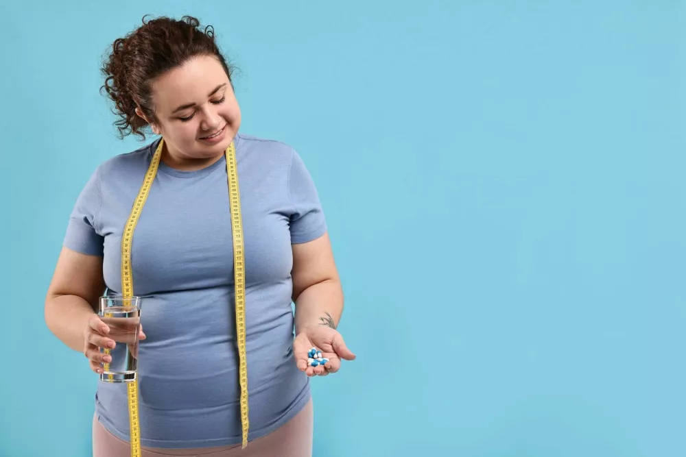 Happy plus size woman holding pile of weight loss supplements and glass with water on light blue background. Space for text