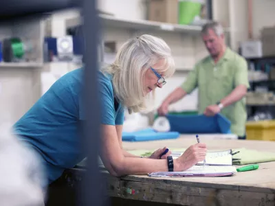 Senior businesswoman is doing paperwork in her stock warehouse. Her husband is in the background measuring fabric.