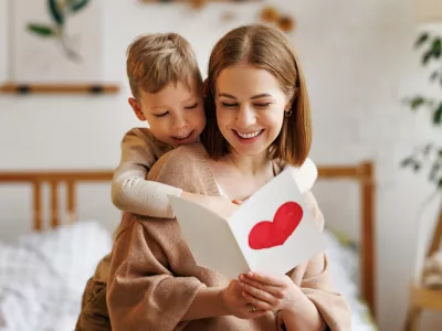 Cute little boy son congratulating his mom happy woman with Mothers day, giving her handmade greeting postcard with red heart while sitting together on bed at home. Family holidays concept / Foto: Evgenyatamanenko