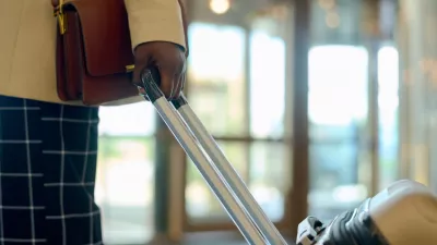 Hand of young black woman in smart casualwear with handbag pulling suitcase with luggage while walking down lounge of hotel