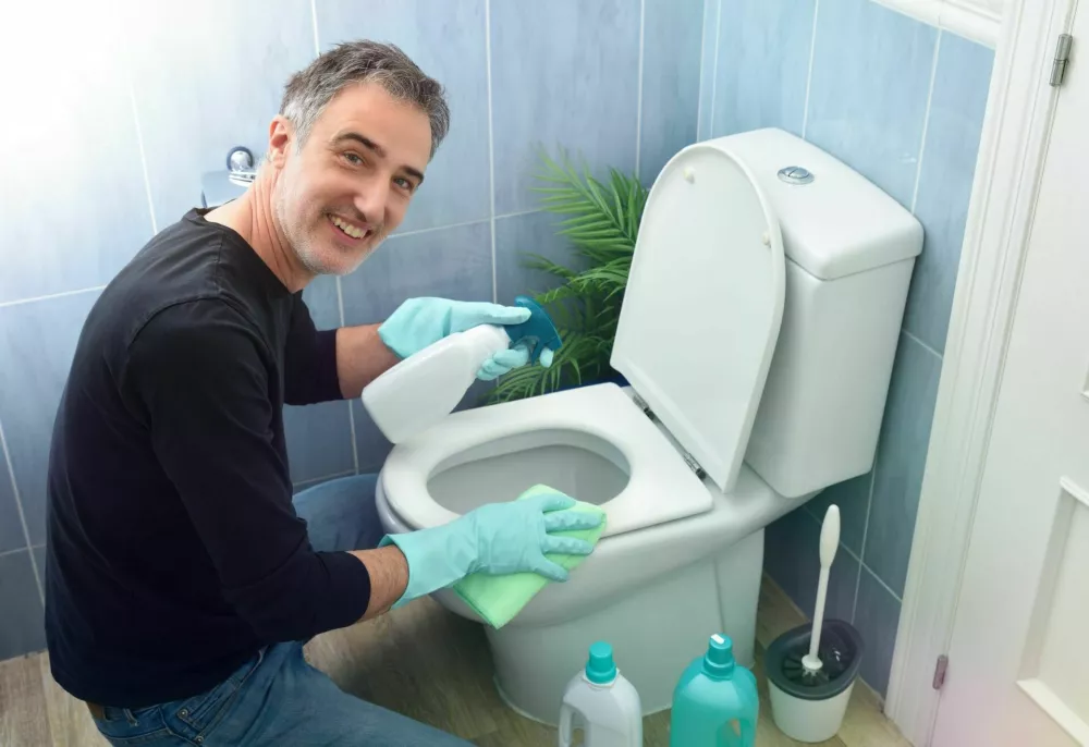 Smiling man looking with green rubber gloves and disinfectant spray bottle scrubbing toilet bowl. Elevated view. / Foto: Davizro