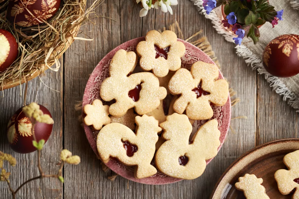 Linzer cookies in the shape of Easter animals filled with strawberry marmalade on a red plate, with decorated eggs / Foto: Madeleine_steinbach