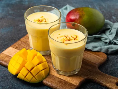 Healthy Indian Ayurveda drink mango lassi in two glasses on rustic concrete table with fresh ripe cut mango, yellow blended beverage made of mango fruit, yoghurt or milk curd and spices / Foto: Thesomegirl