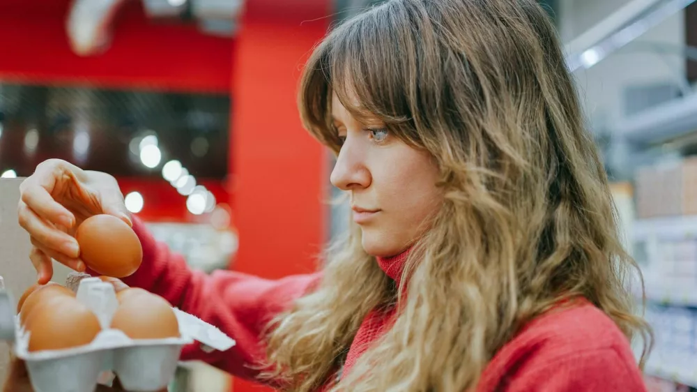 Blonde woman in a red sweater carefully chooses fresh eggs at a modern supermarket, ensuring quality for her nutritious meal