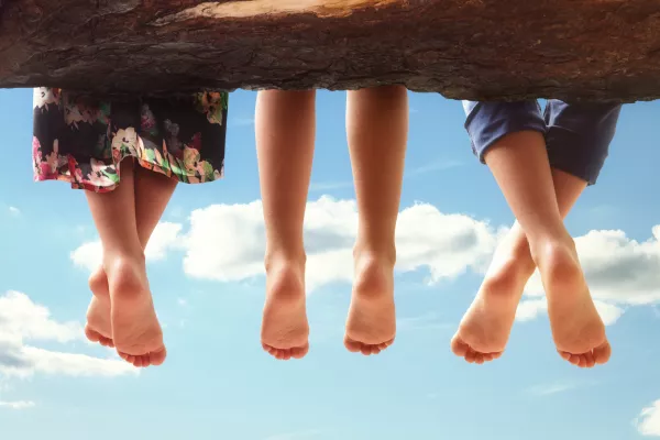 Three kids sitting in a tree dangling their feet against a blue sky in summer concept for family, friends, carefree and vacations