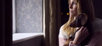 Woman With Pet Dog Trying To Keep Warm By Radiator At Home During Cost Of Living Energy Crisis